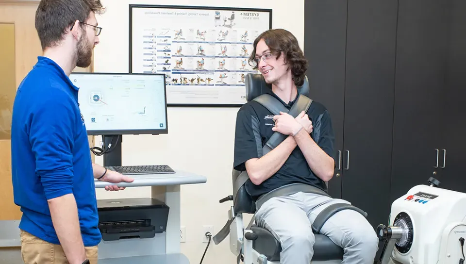 One athletic training student sits upright in a machine that tests leg strength while another student looks at the results on a computer monitor